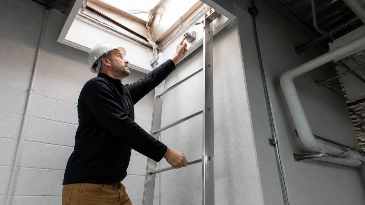 Facility manager using motorized roof hatch with one hand while maintaining secure grip on interior ladder with other hand, demonstrating safe one-handed operation in commercial building interior