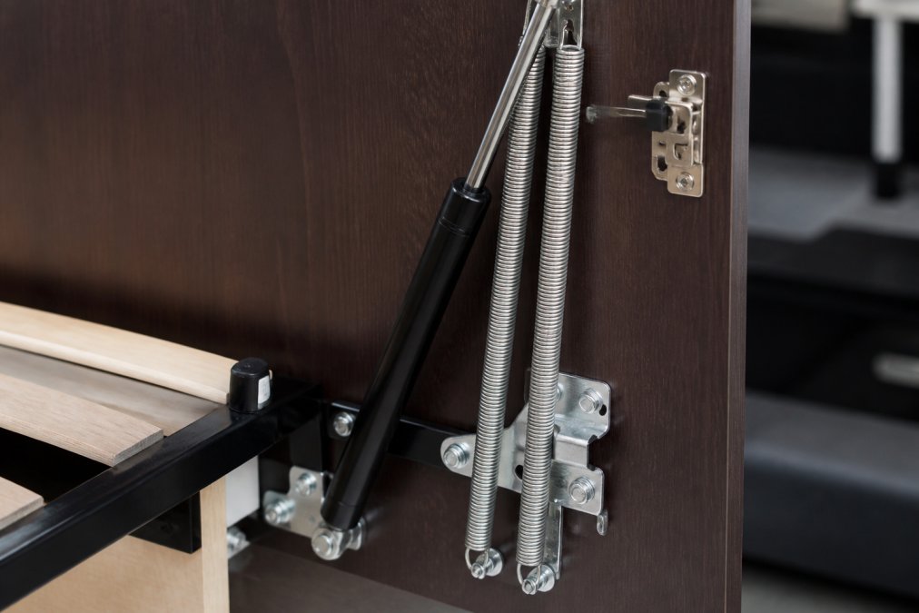 Close-up of a Murphy bed piston mechanism and safety latch hardware showing counterbalance springs and metal fasteners in detail, with professional lighting highlighting the mechanical components