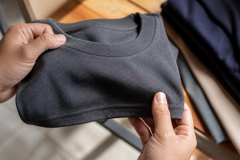 Close-up of hands holding the collar and hem of a folded graphic t-shirt, showing the fabric texture and stitching detail under bright natural light