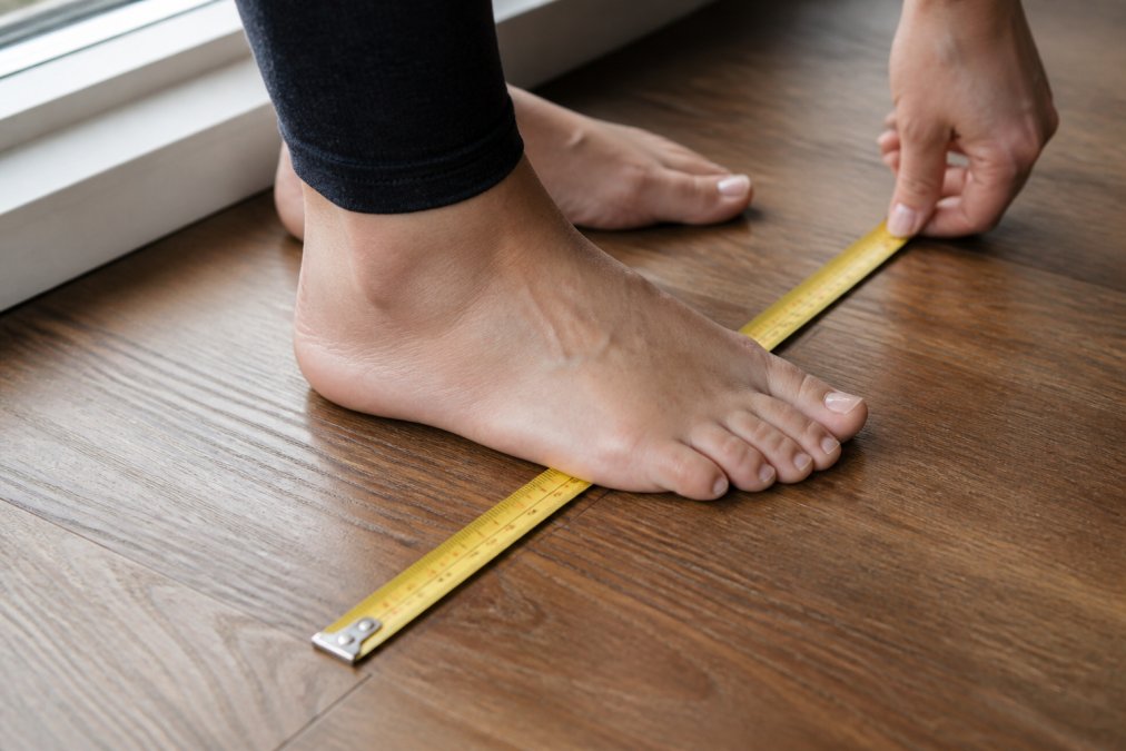 Close-up of feet positioned on a ruler or measuring tape on a hardwood floor, showing proper heel-to-toe measurement technique with natural window lighting