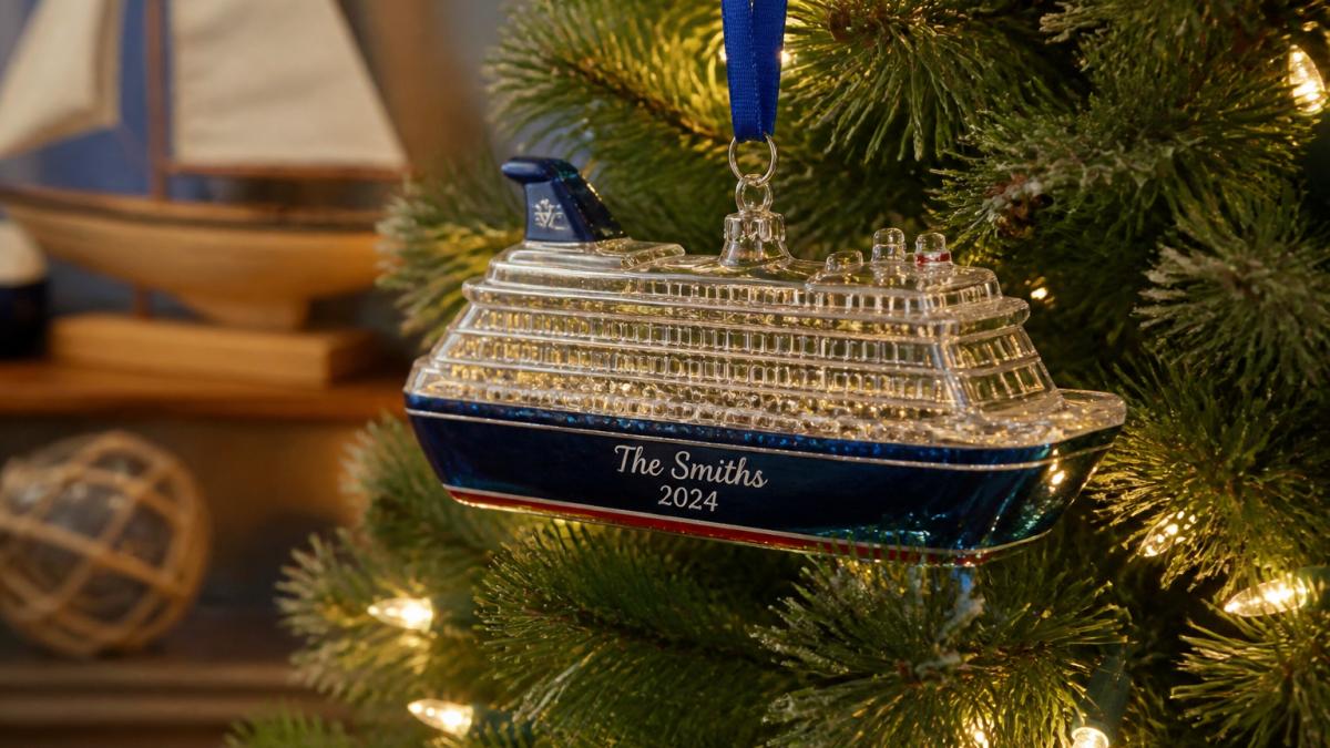 Close-up of a hand-personalized glass cruise ship ornament hanging on a lit Christmas tree, with a small name and date visible on the ornament