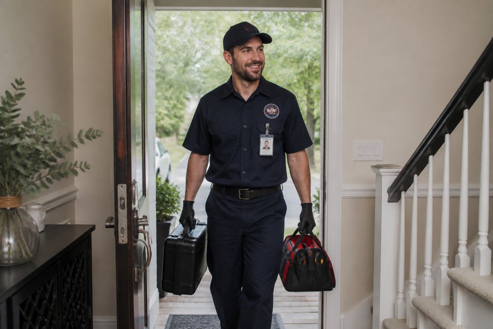 Professional pest control technician in clearly visible company uniform with branded logo, ID badge displayed prominently, and carrying professional equipment cases entering a residential home through front door