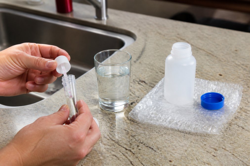 Hands holding a water testing kit over a sink to check if are pfas in my city water with a lab sample bottle.