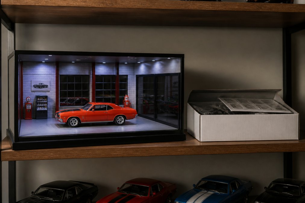 A collector's home display shelf showing a 1:18 scale diecast muscle car inside an illuminated pre-built dealership diorama, with a second unassembled diorama kit box resting beside it