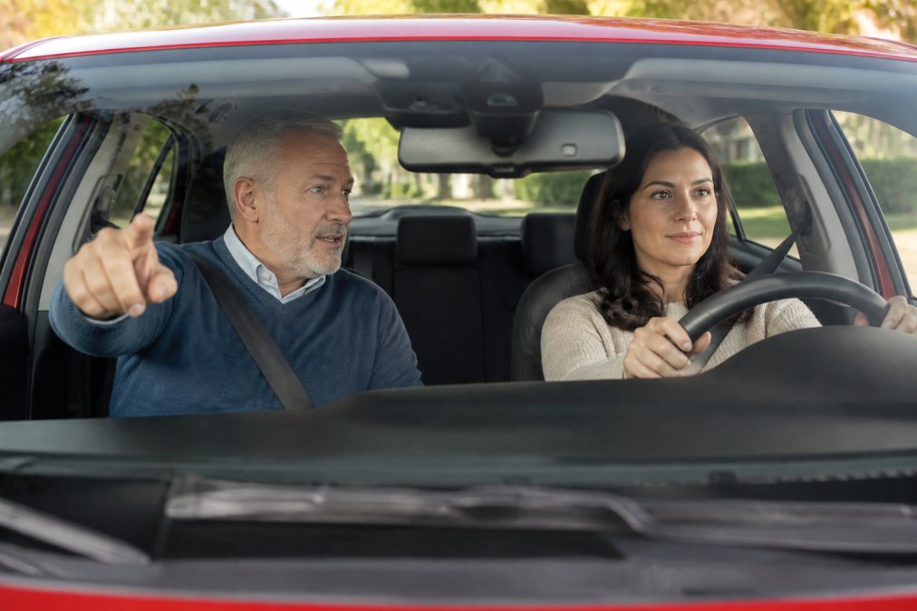 A calm, professional driving instructor sitting in the passenger seat of a car, smiling and giving directions to a relaxed adult student driver in a suburban neighborhood