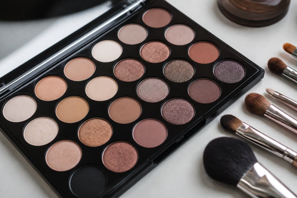 Close-up of an open professional eyeshadow palette with richly pigmented neutral and shimmer shades, laid flat on a clean white surface with a few makeup brushes resting beside it