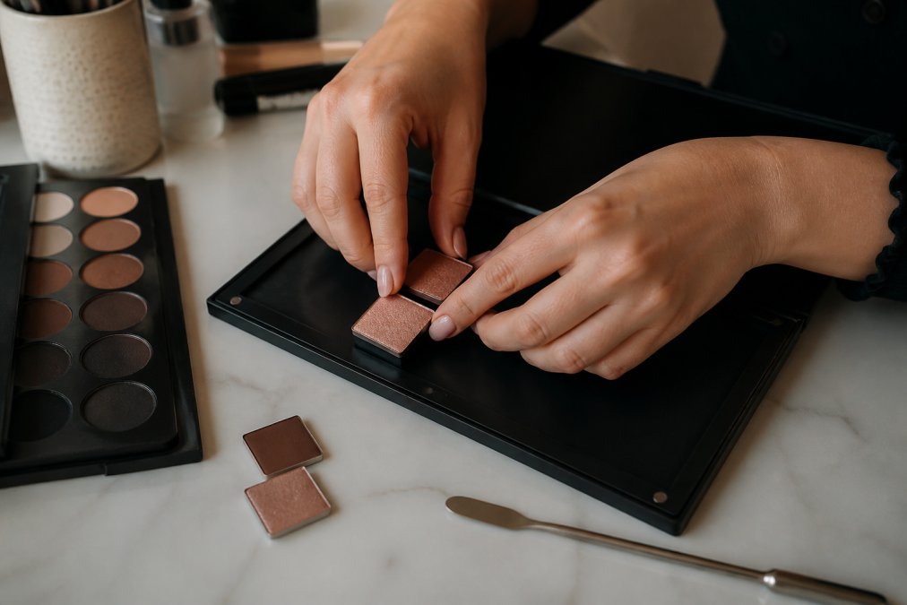 Hands carefully pressing individual eyeshadow pans into a magnetic palette organiser on a tidy dressing table, with a spatula and a few loose pans visible nearby