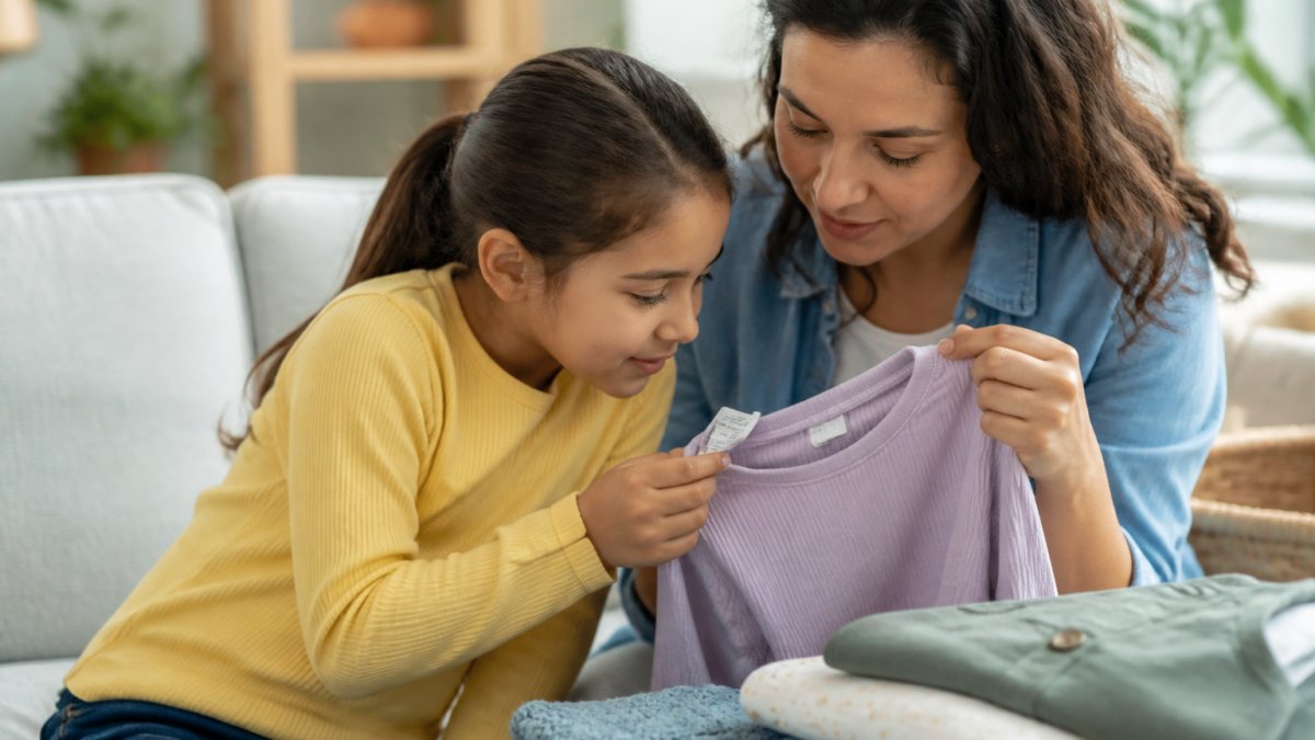 Parent and child examining clothing labels and tags together in a bright home setting, reading fabric content information on a shirt