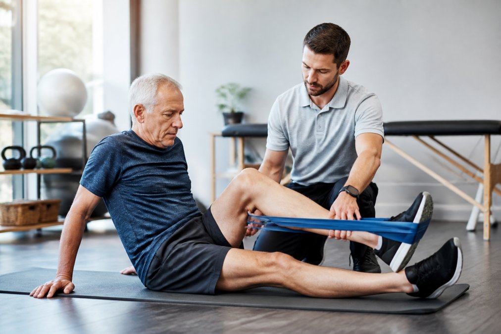 A patient in their 60s performing guided knee rehabilitation exercises with a physical therapist in a bright, modern clinic, resistance band and exercise mat visible