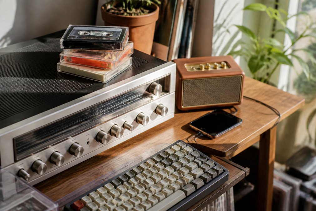 A sunlit living room shelf holding a vintage stereo receiver, a stack of cassette tapes, a mechanical keyboard, and a modern smartphone playing music through a small retro speaker
