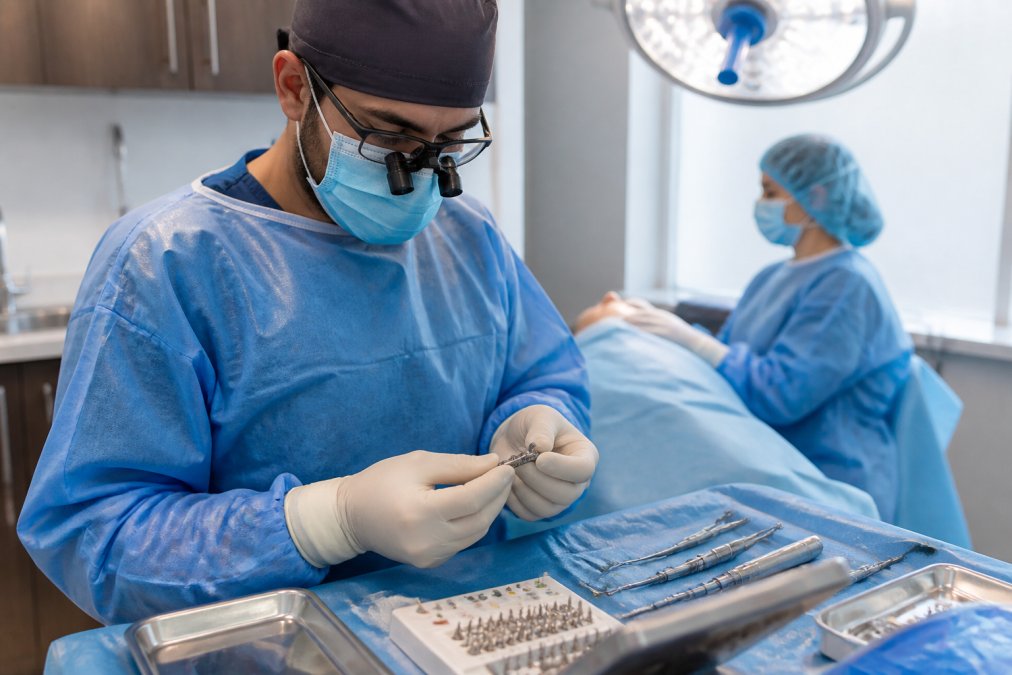 Dental surgeon in surgical attire and magnification loupes preparing sterile titanium implant components and surgical instruments on a draped operative tray in a modern operating room with overhead surgical lighting