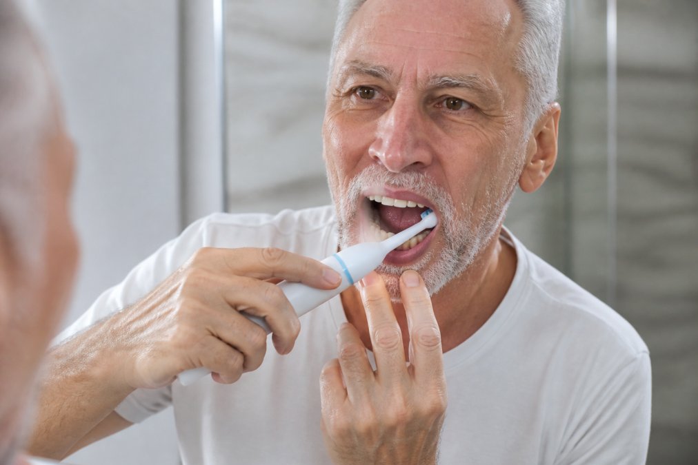 Close-up of a person's hands demonstrating proper electric toothbrush technique along the gum line around the front lower teeth, with soft bristles angled at 45 degrees toward the gum tissue, in natural bathroom lighting
