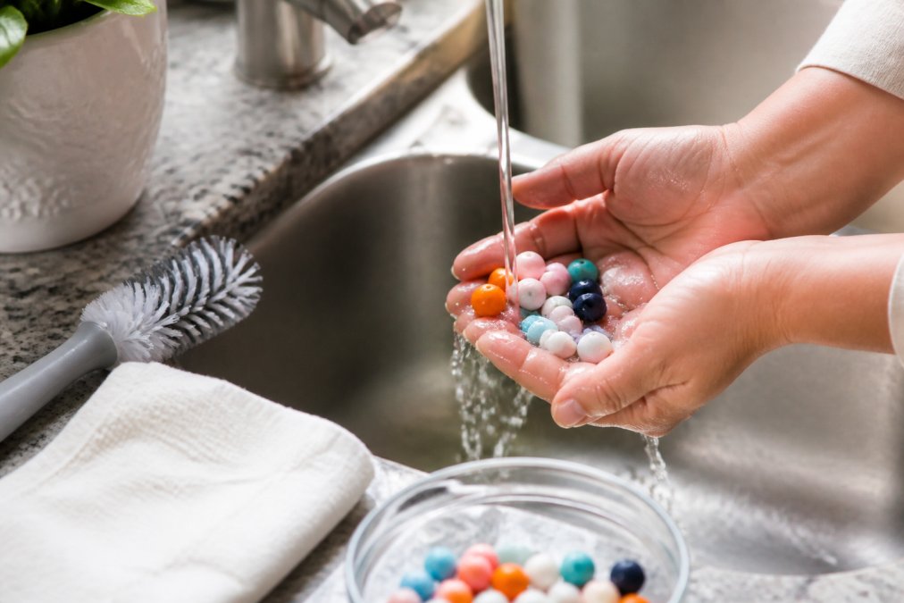 Close-up of hands washing colorful silicone beads under warm running water in a sink, with a soft brush nearby and a clean cloth ready for drying