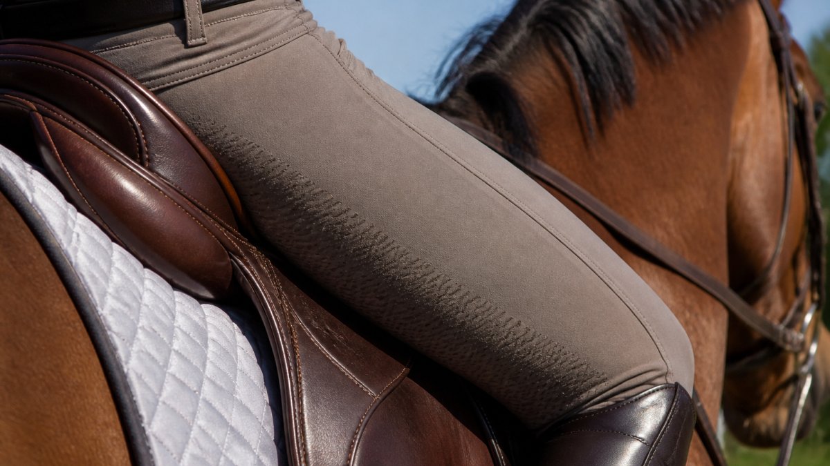 Close-up of a rider's leg and lower body in silicone grip breeches while seated in a saddle, showing the contact point between the thigh and saddle leather during a ride, with natural outdoor lighting