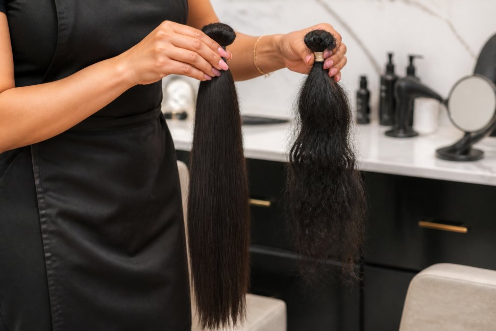 Close-up of a hair extension specialist's hands holding two bundles of raw hair side by side, one sleek and aligned, the other tangled, in a bright salon setting