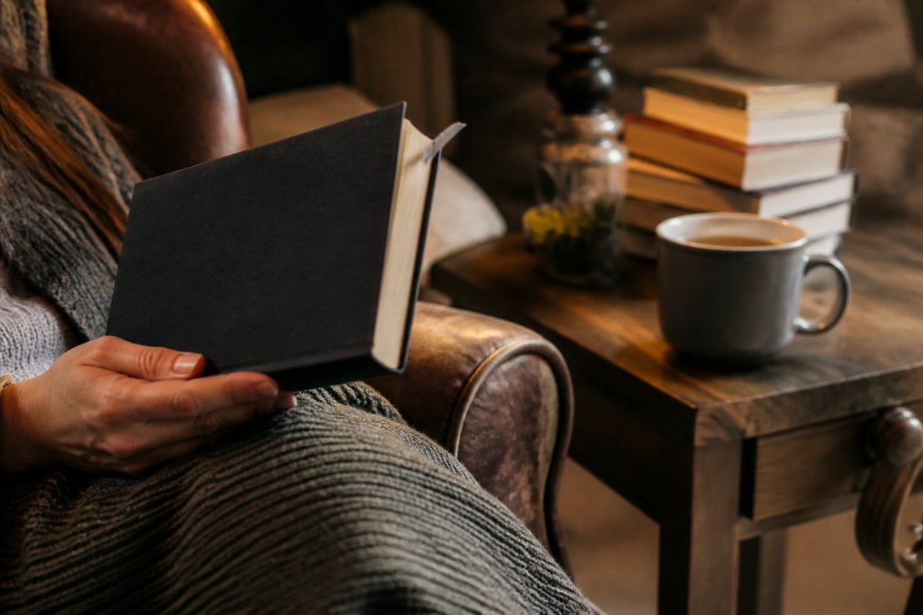 Hands holding a book with a bookmark in a cozy armchair with tea, perfect for reading slow burn thrillers.