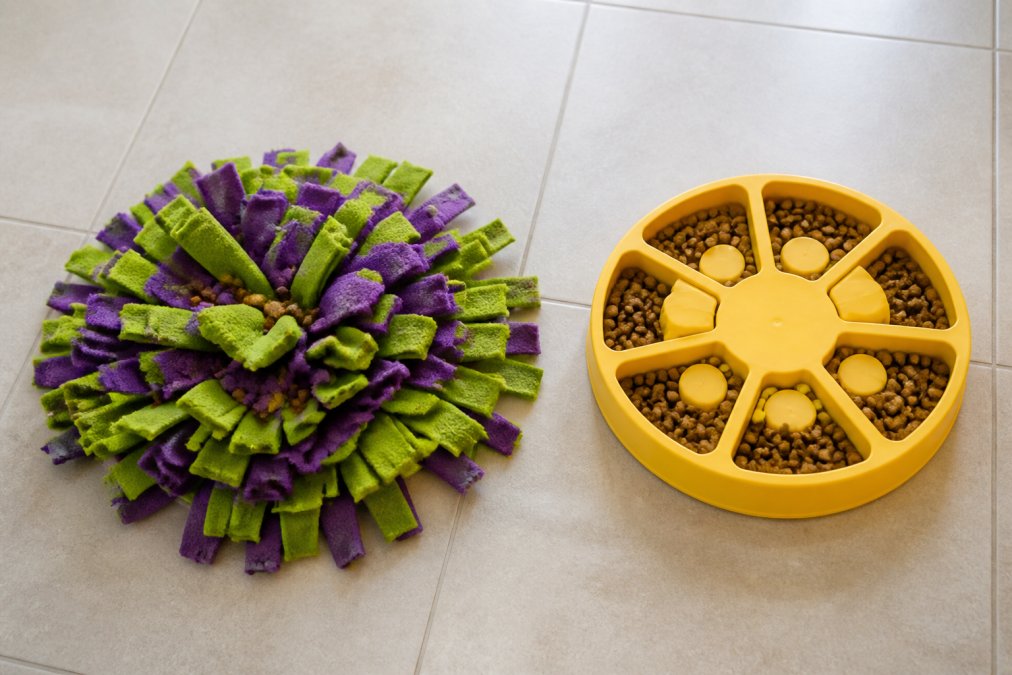 Side-by-side view on a light-colored kitchen tile floor: a green and purple fleece snuffle mat with small treat pieces tucked into the strips on the left, and a yellow plastic interactive puzzle feeder bowl with food visible in the compartments on the right, both photographed from above in bright indoor lighting