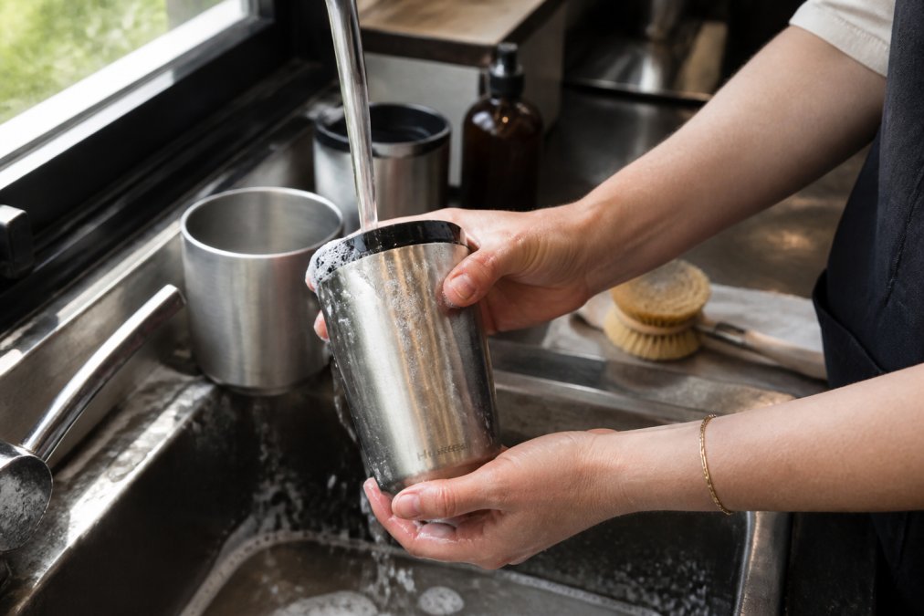 Hands rinsing a stainless steel cup under warm running water at a cafe sink, demonstrating proper cleaning technique with soap suds visible, natural light from the window