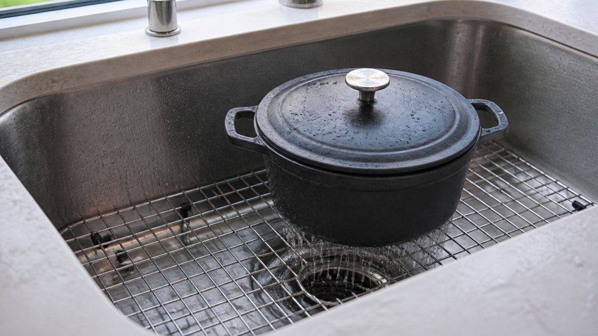 Stainless steel pot resting on a metal sink grid as water drains underneath in a sunlit kitchen basin