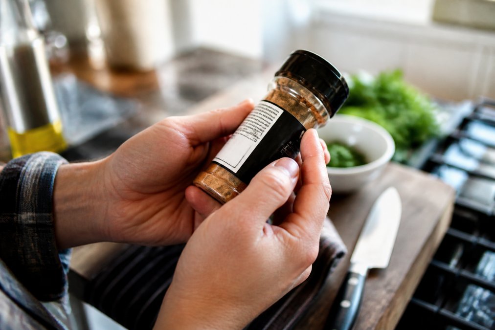 Close-up of hands holding a spice jar, carefully examining the ingredient list and allergen statement printed on the back label in bright kitchen lighting
