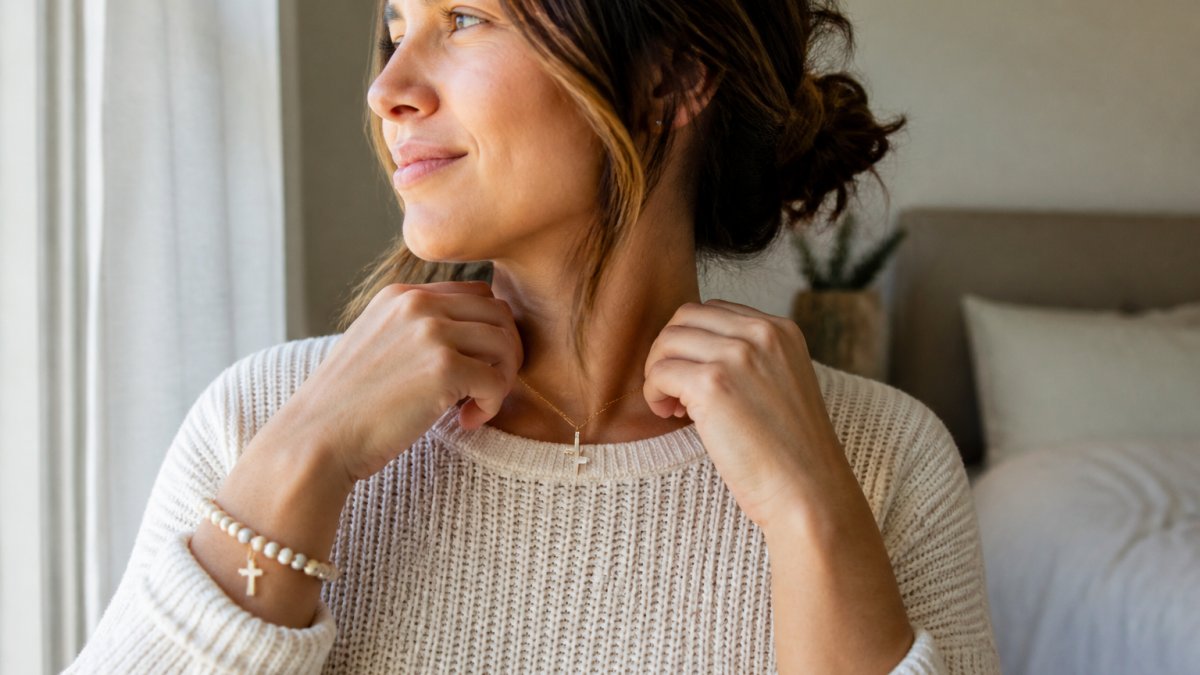 A woman in a cream knit sweater fastening a delicate cross necklace at her collarbone, a beaded faith bracelet on her wrist, soft morning window light in a calm bedroom