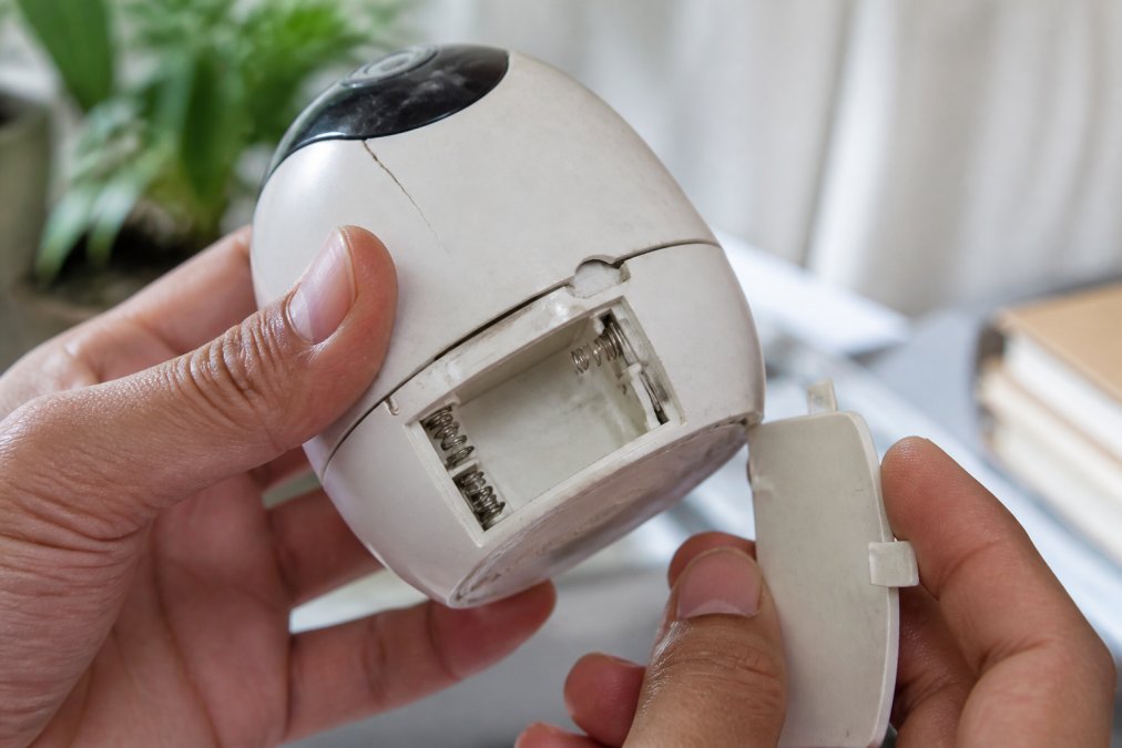 Close-up of hands holding a smart home device with visible wear, loose battery compartment, and small cracks in plastic casing under natural light