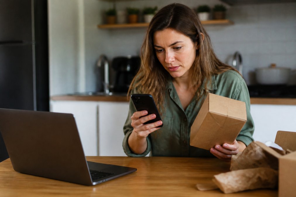 A shopper holding a smartphone and examining a newly delivered product box at their kitchen table, with a laptop showing a product page nearby
