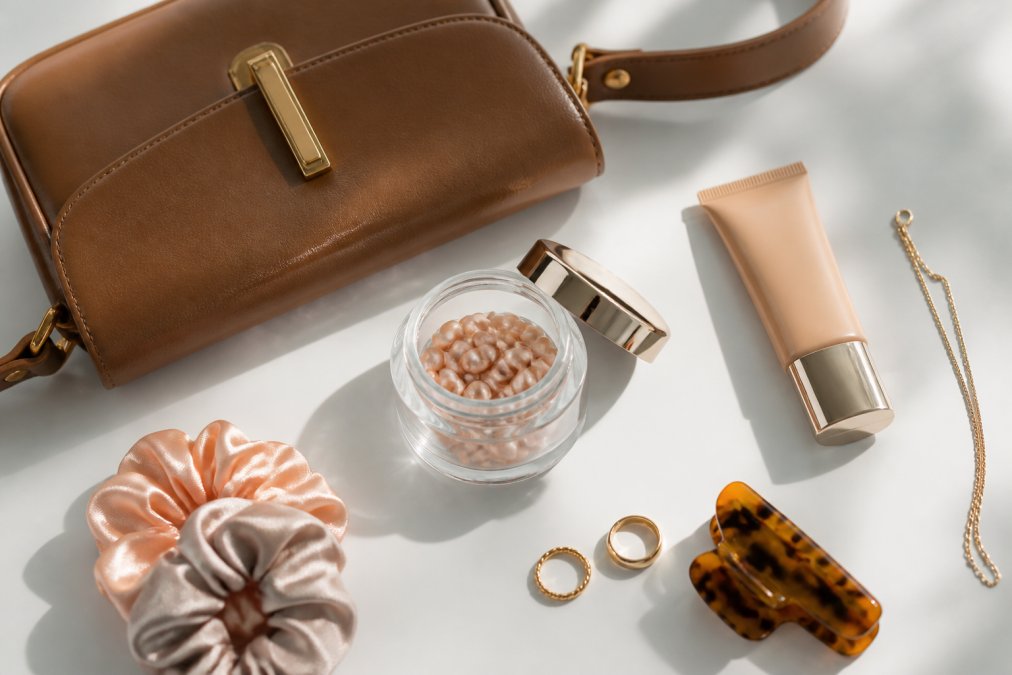 Flat lay of several trending lifestyle and beauty products arranged on a clean white surface, including a stylish leather handbag, a jar of skincare capsules, and a CC cream tube, photographed from directly above in bright natural light