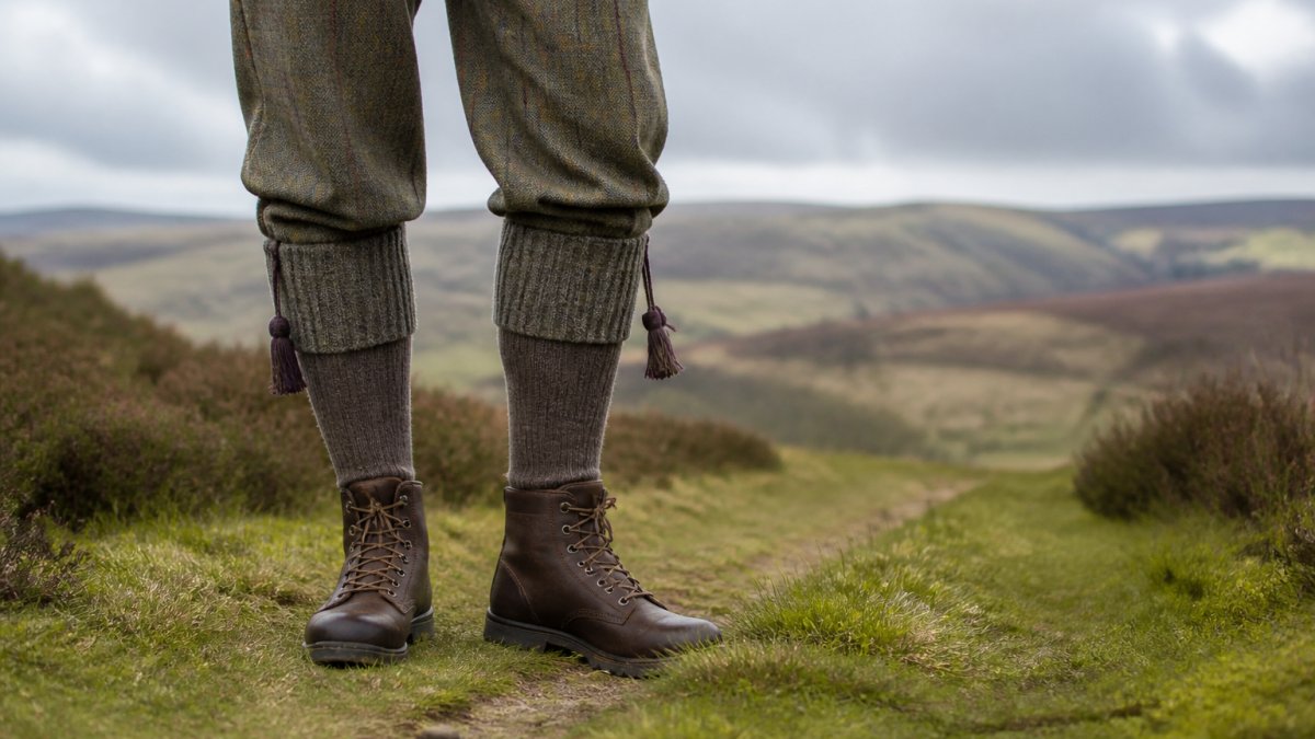 Close-up of a person's lower legs wearing woollen shooting socks turned over at the calf with garter ties, worn with tweed breeks and country boots, standing on a grassy moorland track