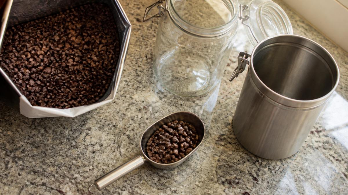 Overhead shot of a kitchen counter with an open bag of whole roasted coffee beans, a glass canister with a clamp lid, a stainless steel container, and a scoop, natural window light