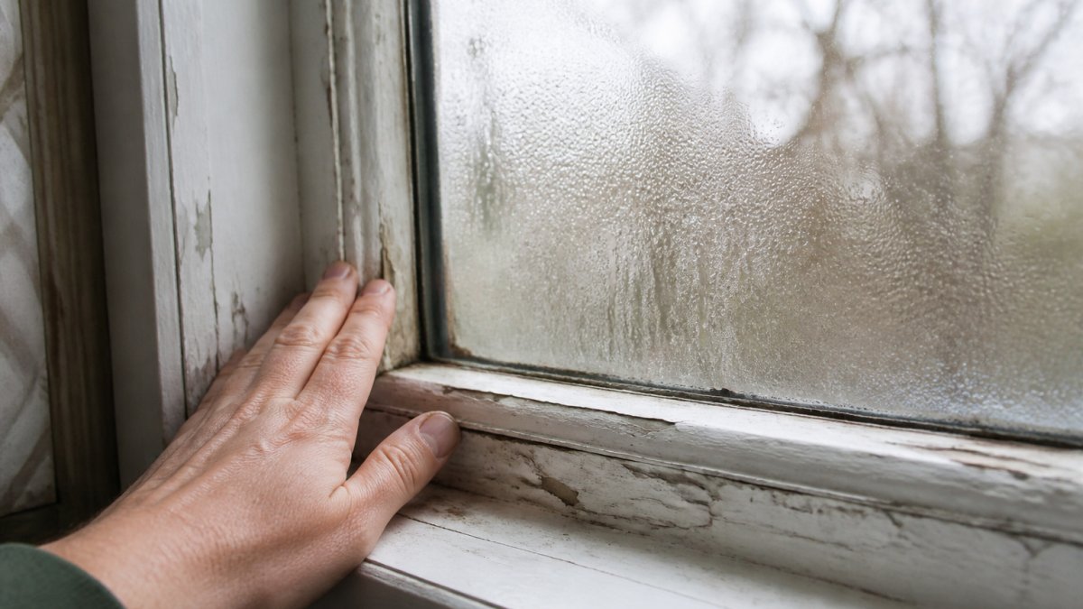 Close-up of a homeowner's hand running along a drafty window sash with visible condensation and peeling paint on an older wooden frame