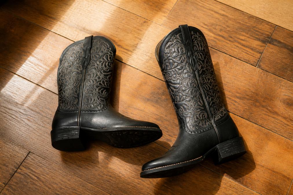 Close-up of a pair of black leather cowboy boots with western stitching detail laid flat on a warm-toned wooden floor, natural light casting soft shadows