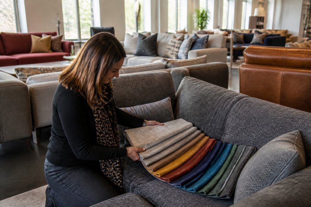 Customer examining fabric samples and upholstery options on display sofas in a bright, elegant showroom with natural light filtering through tall windows