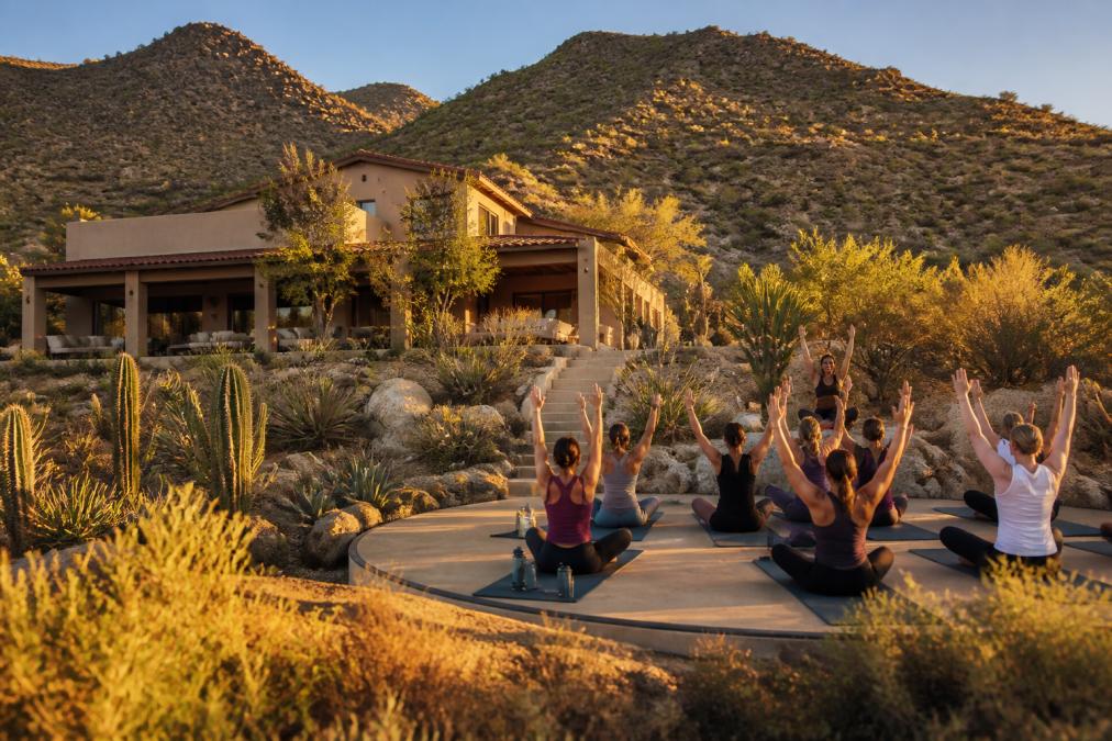 Serene desert sanctuary at golden hour with a modern wellness villa nestled against Sunrise Mountain, desert vegetation in foreground, peaceful outdoor yoga space visible