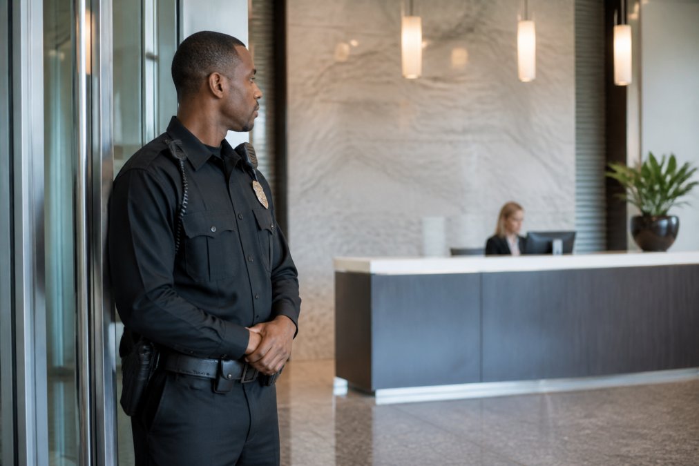 A professional security guard in uniform standing at a modern office building lobby entrance, monitoring the front desk area with a calm, alert posture