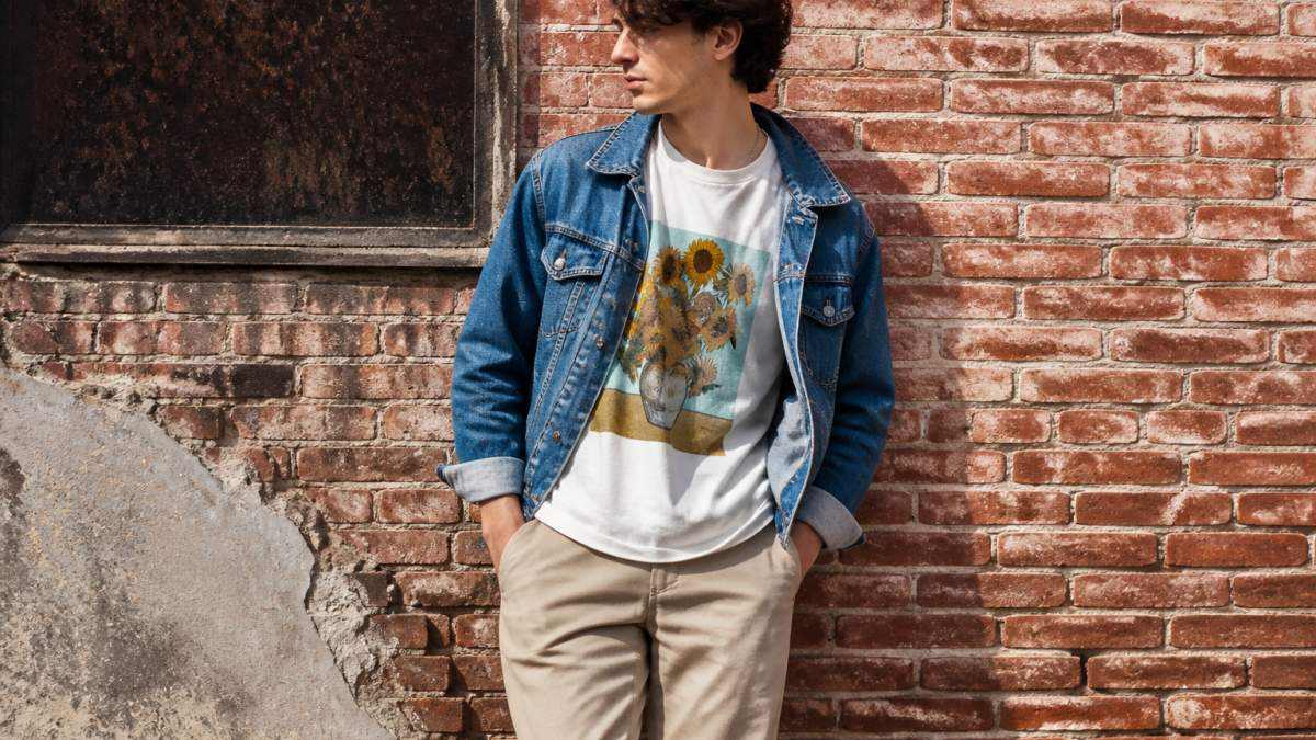 Person wearing a vintage-washed art-inspired tee layered with a denim jacket and neutral pants, standing in natural outdoor lighting against a textured brick wall