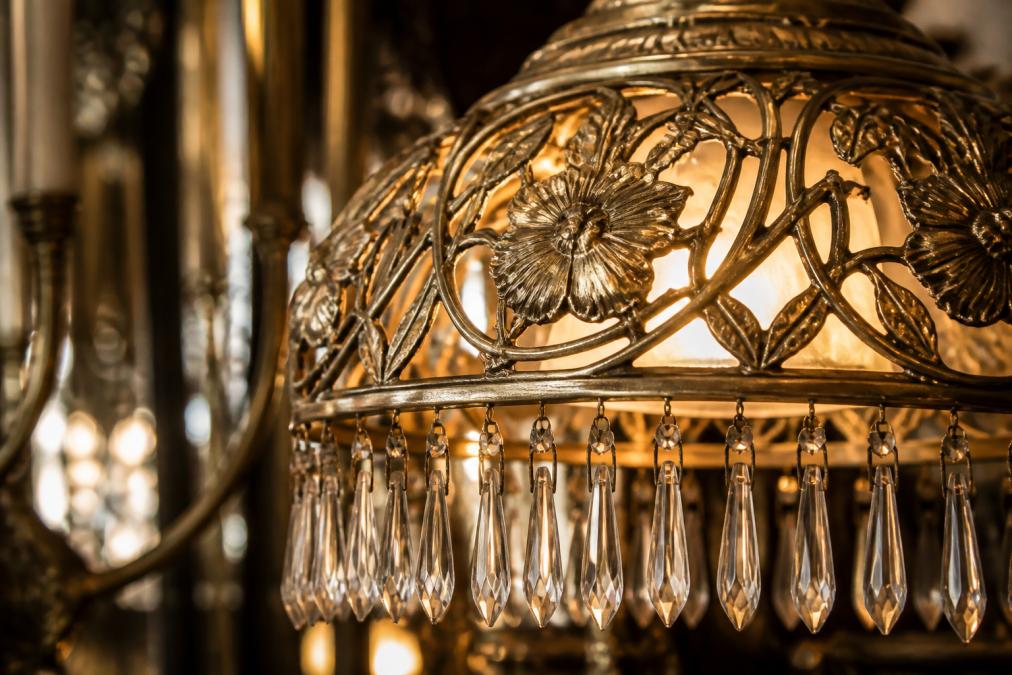 Elegante Nahaufnahme einer handgefertigten Messinglampe im Jugendstil mit filigranen floralen Ornamenten in Reliefform, Kristallelemente brechen das warme Licht, detailreiche Metallverzierungen sichtbar