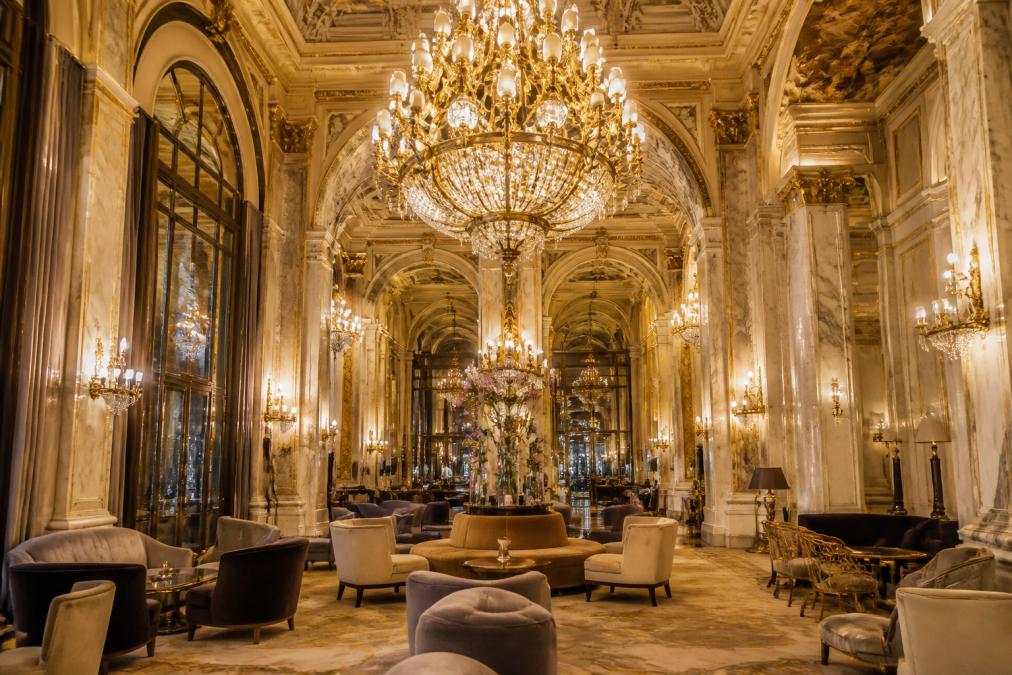 Luxuriöse Hotellobby mit imposantem Art Nouveau Kronleuchter aus Messing und Kristall, umgeben von eleganten Möbeln, klassischer Architektur, warmes Licht reflektiert in Marmor und Spiegeln