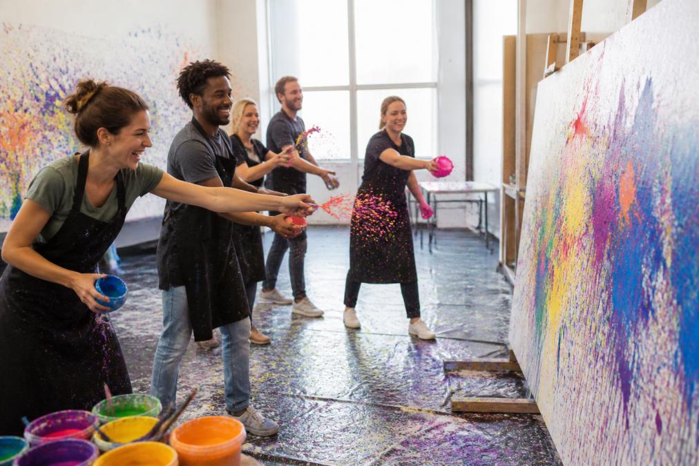 Gruppe von Kolleginnen und Kollegen in legerer Businesskleidung lacht gemeinsam, während sie Farbe auf eine große Leinwand in einem hellen, geräumigen Studio mit bunten Spritzern auf Boden und Wänden wirft