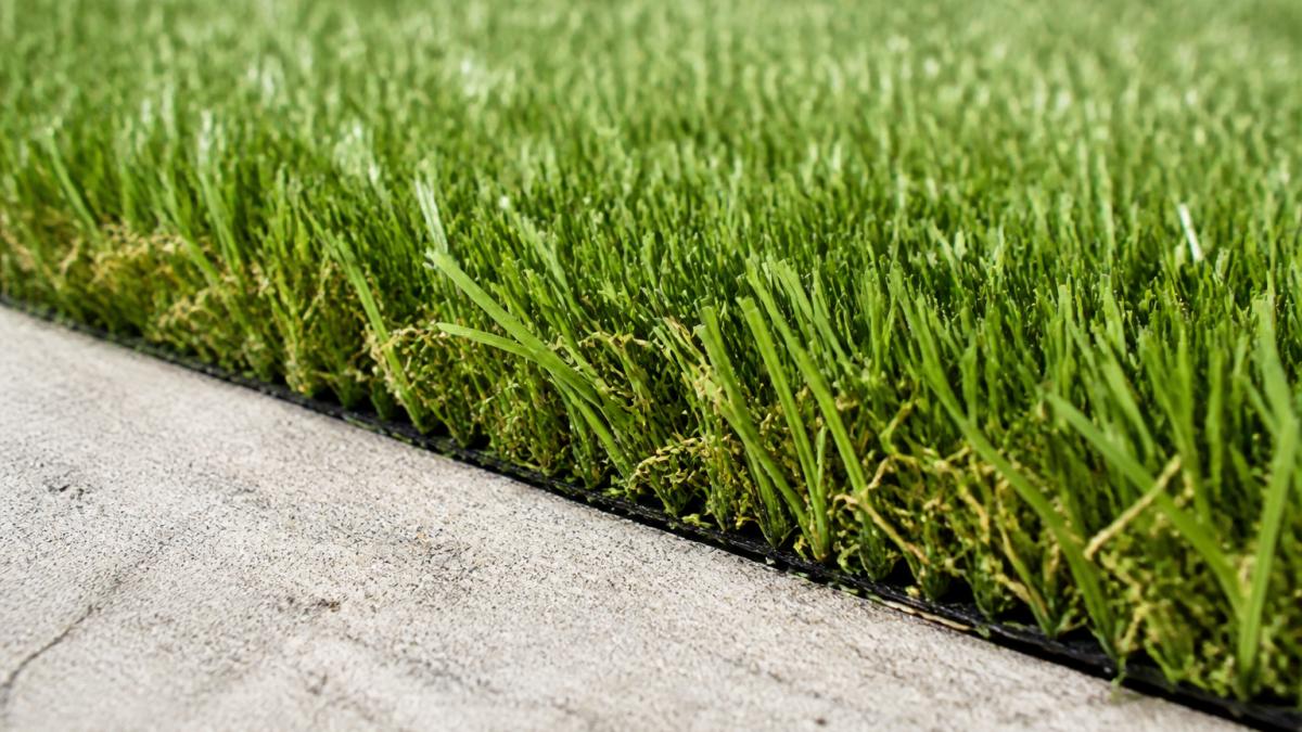 Close-up of freshly installed artificial grass on concrete patio showing multi-color polyethylene fibers with natural grass appearance, concrete edge visible, bright daylight