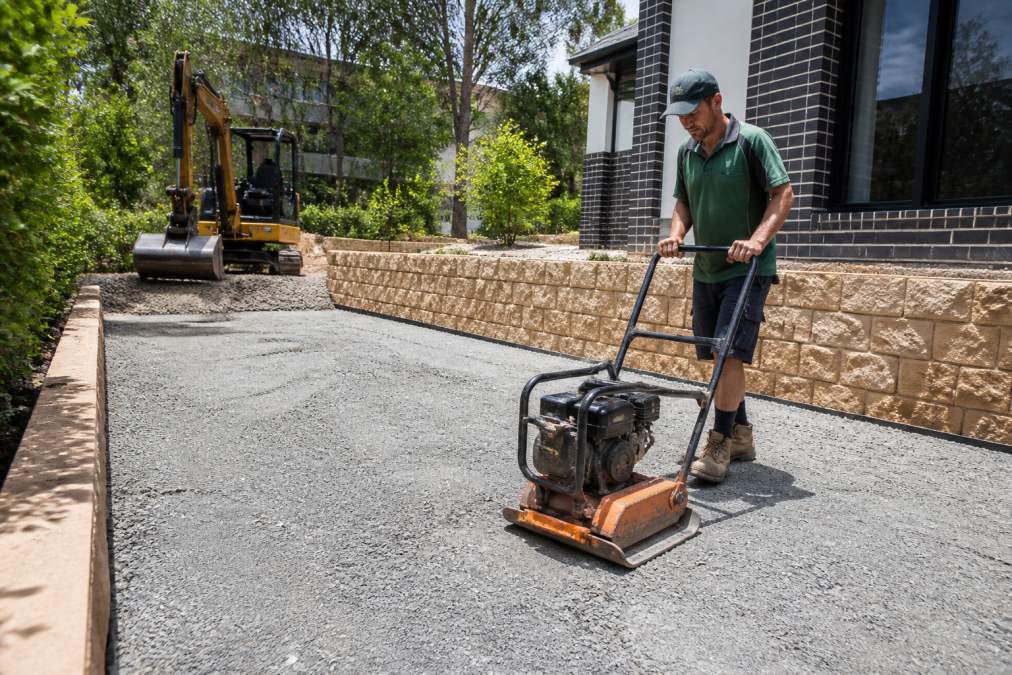 Professional landscaper preparing and compacting crushed rock base layer on residential property in North-West Sydney, with excavator visible in background and proper grading in place