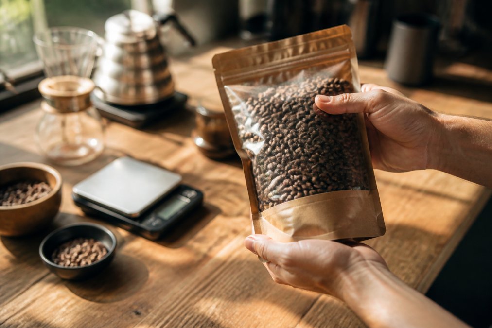 Close-up of freshly roasted coffee beans with visible roast date on label, held over wooden surface with coffee brewing equipment and scales in soft morning light