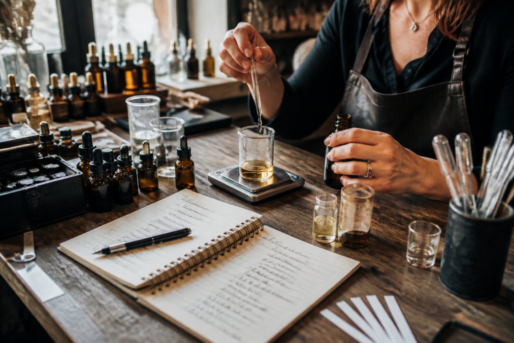 Artisan perfumer carefully measuring fragrance oils using glass pipettes and precise instruments, with handwritten formula notes and ingredient labels visible on wooden workbench with warm studio lighting