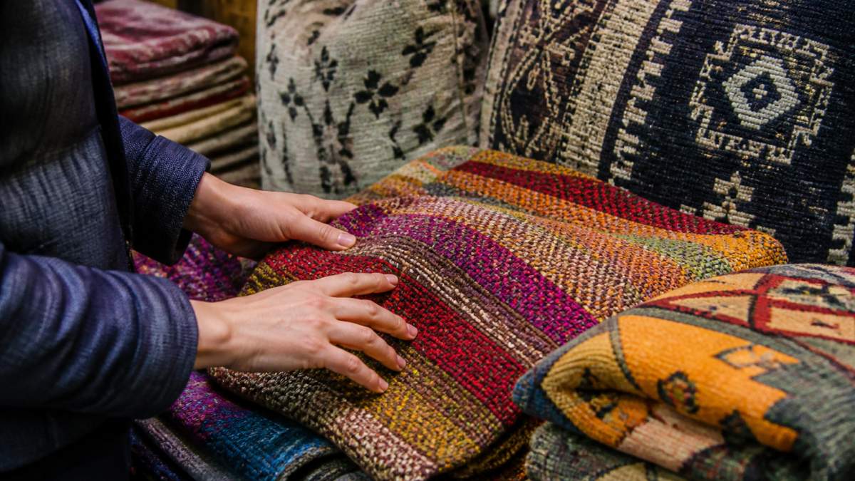 Close-up of hands touching colorful handwoven textiles and patterned pillows displayed together, showing intricate weave patterns and fabric texture details