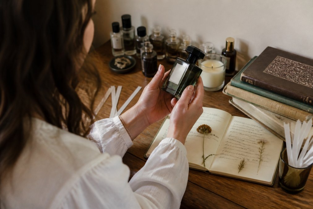Fragrance enthusiast holding a luxury artisan perfume bottle, carefully examining the label and packaging details under warm natural light, with vintage perfume books and scent notes visible on the table beside them