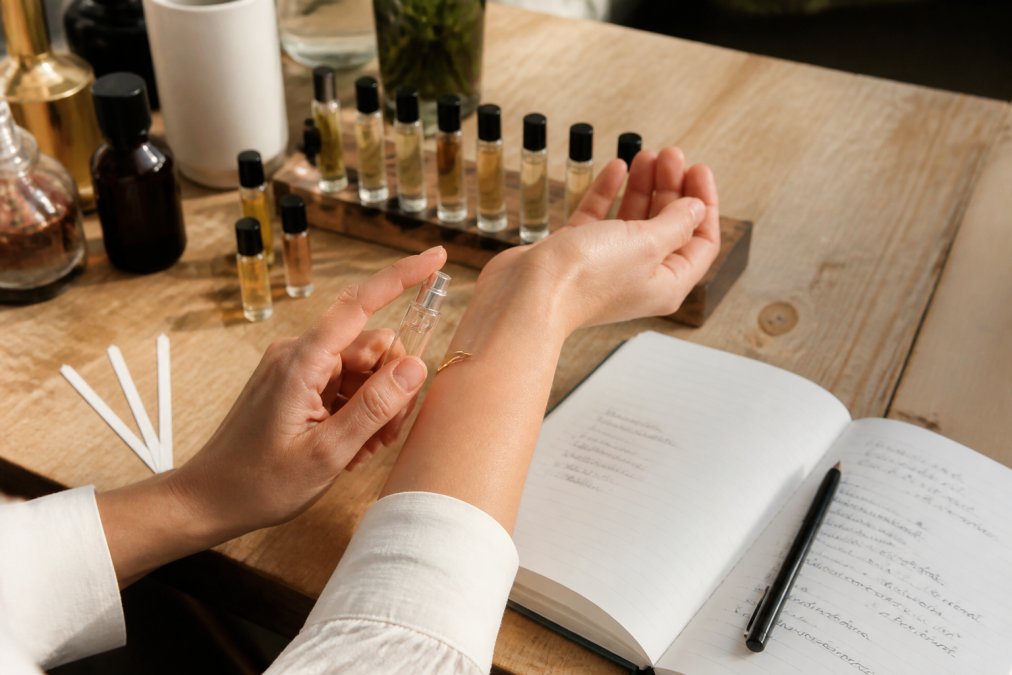 Close-up of someone's hands testing fragrance samples on their inner wrist, with multiple sample vials arranged on a wooden surface and an open scent journal with handwritten notes nearby