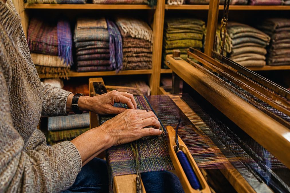 Weaver's hands working at a traditional pedal-powered loom, fingers guiding colourful wool threads through the loom, with finished woven scarves and throws stacked on shelves in the studio background