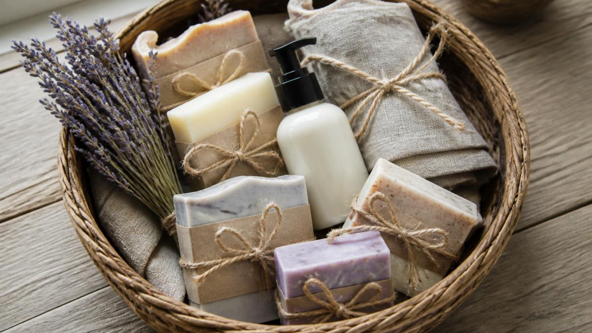 Overhead photo of a woven basket filled with wrapped handmade soap bars, a small bottle of lotion, dried lavender sprigs, and a folded linen cloth on a rustic wooden table in soft window light