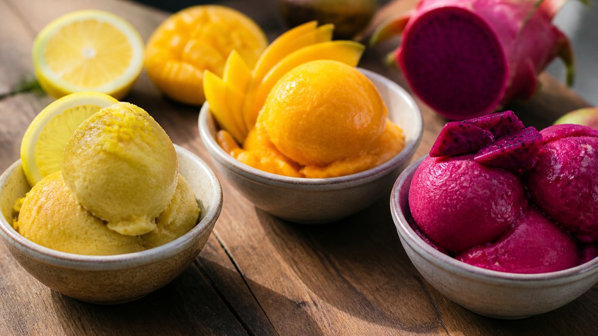 Close-up of vibrant artisan sorbet scoops in serving bowls, showcasing rich yuzu yellow, deep mango orange, and striking dragon fruit pink with fresh fruit garnish and natural daylight