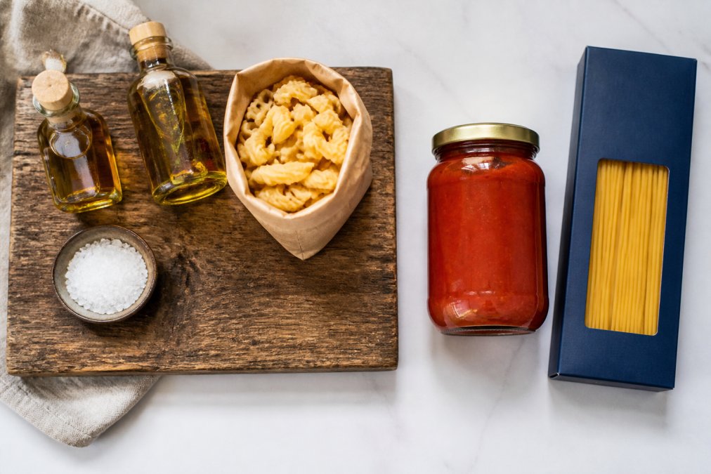 Overhead shot of a rustic wooden board holding small-batch olive oil bottles, bronze-cut pasta, and flaky sea salt in kraft paper packaging, set beside a clean white counter with a jar of commercial pasta sauce and a box of standard pasta